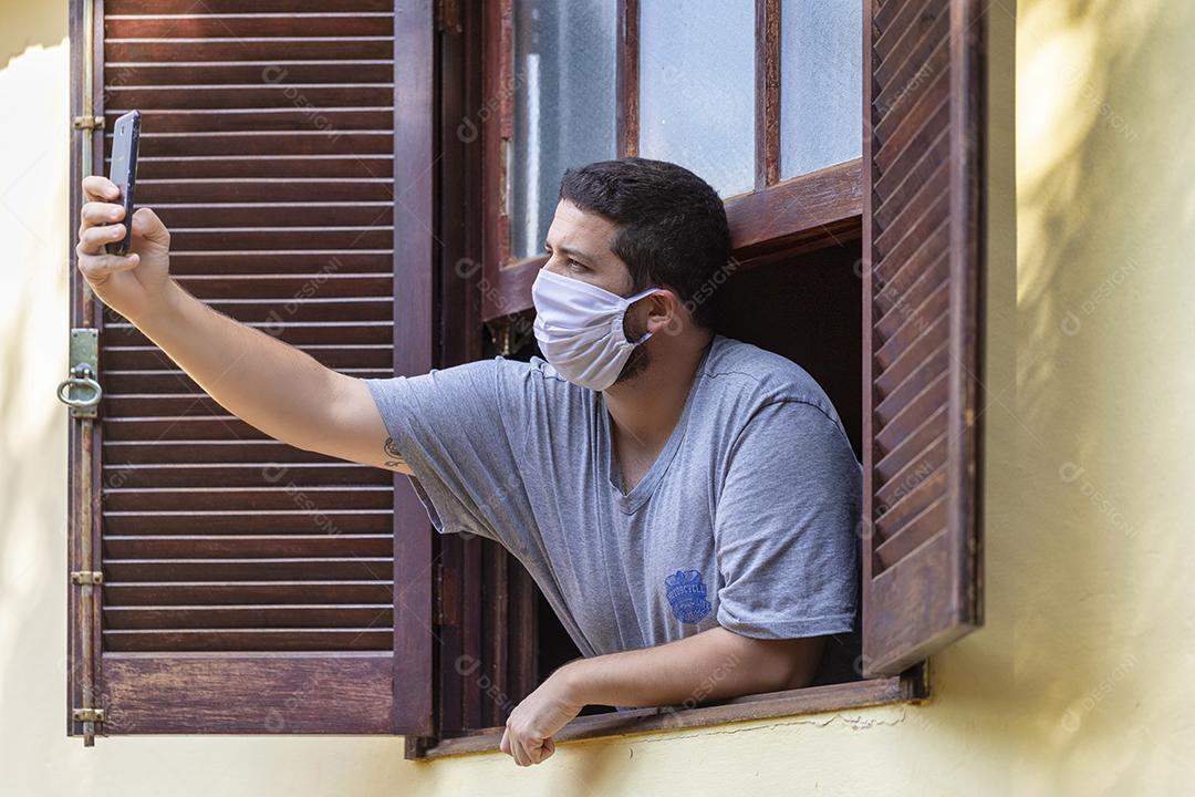 homem com máscara de proteção na janela de casa tirando self