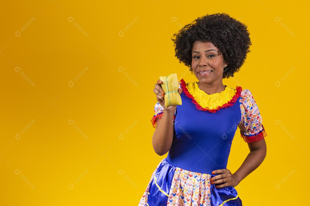 mulher afro em roupas de festa junina segurando um tamale em fundo amarelo