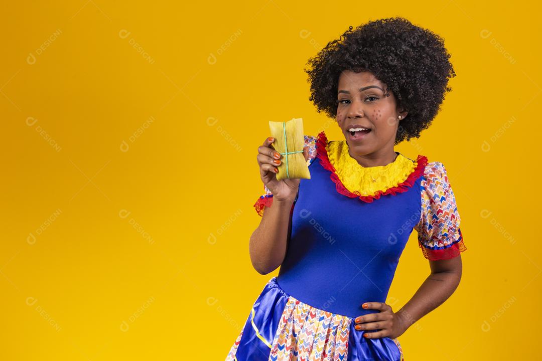 mulher afro em roupas de festa junina segurando um tamale em fundo amarelo