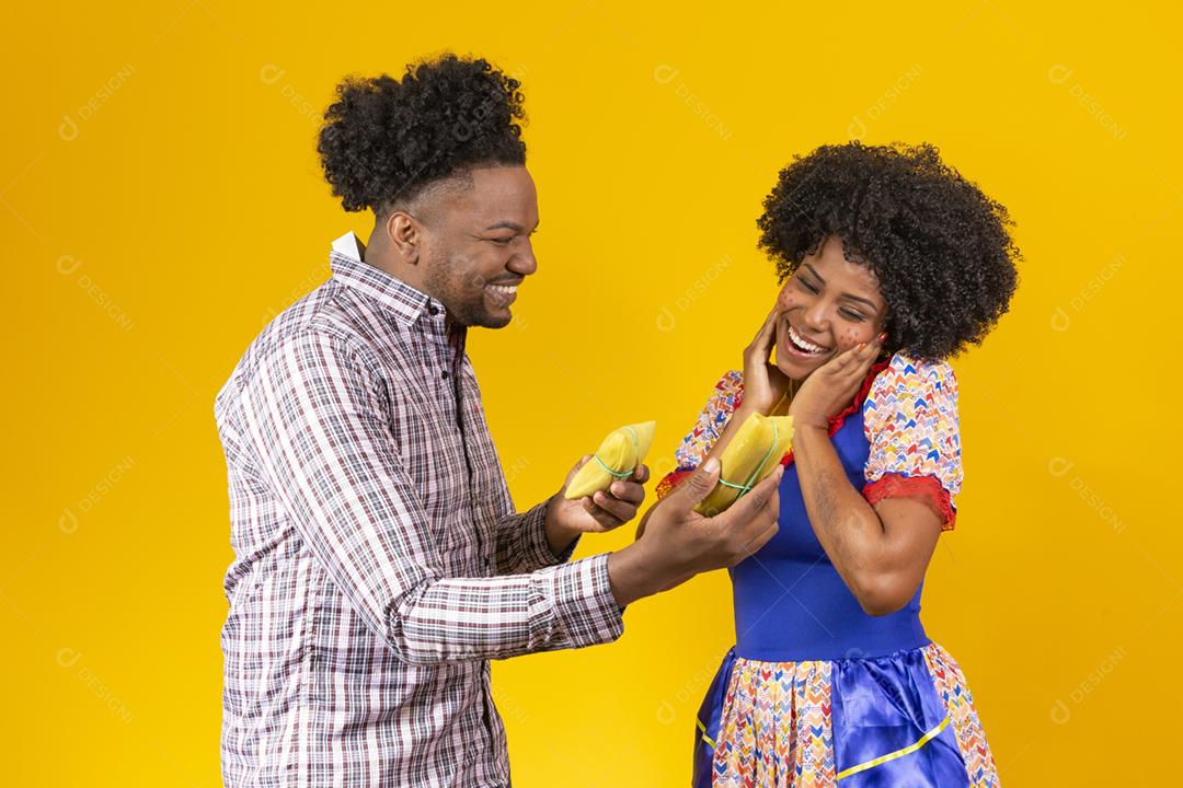 casal de namorados afro em roupas de festa junina segurando uma pamonha
