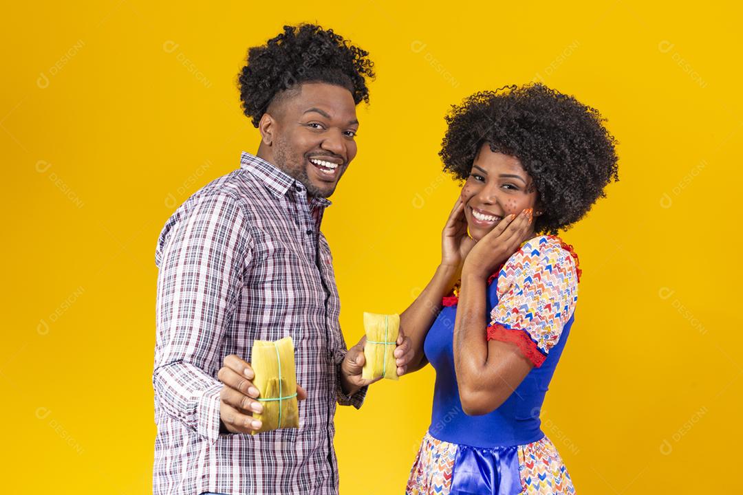 casal de namorados afro em roupas de festa junina segurando uma pamonha