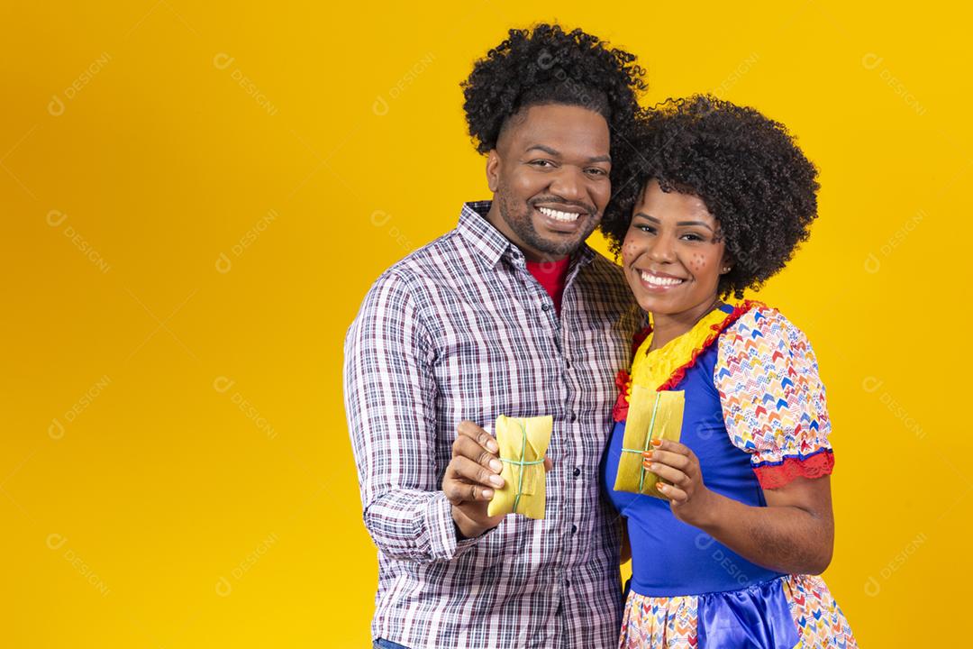 casal de namorados afro em roupas de festa junina segurando uma pamonha