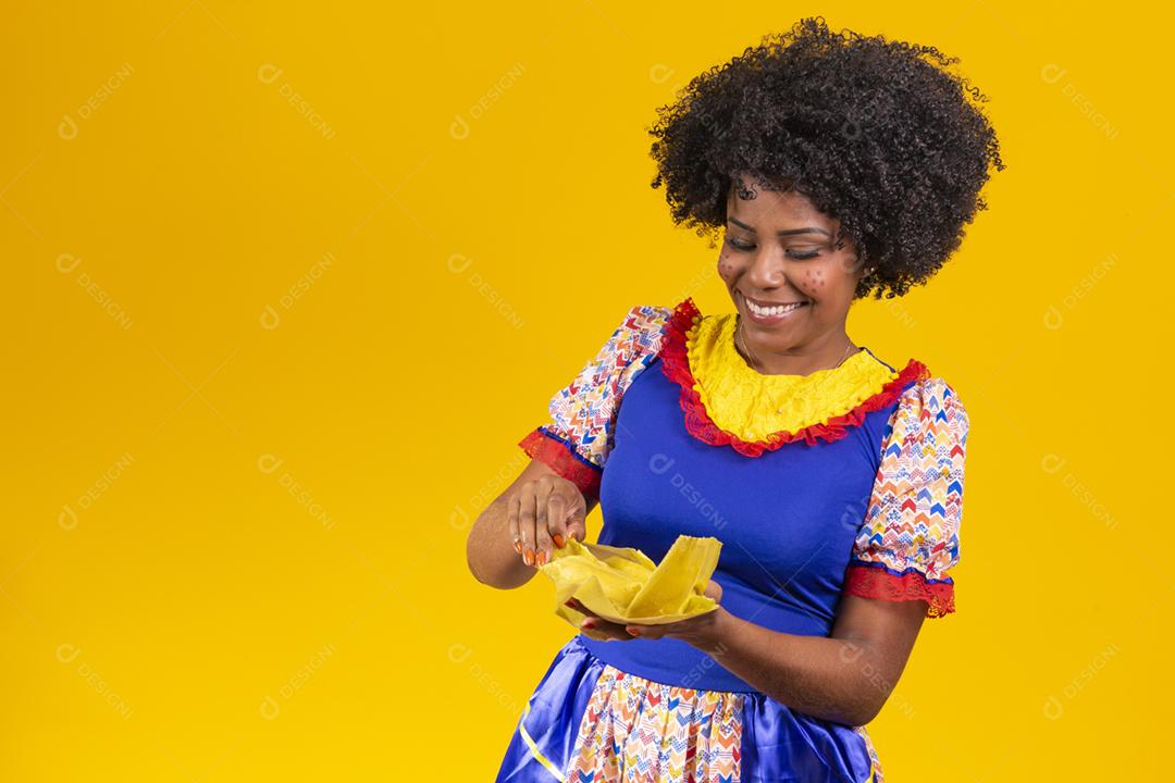 Mulher negra com roupas típicas da festa junina comendo uma deliciosa pamonha