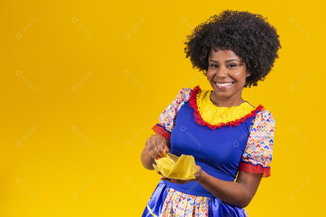 Mulher negra com roupas típicas da festa junina comendo uma deliciosa pamonha