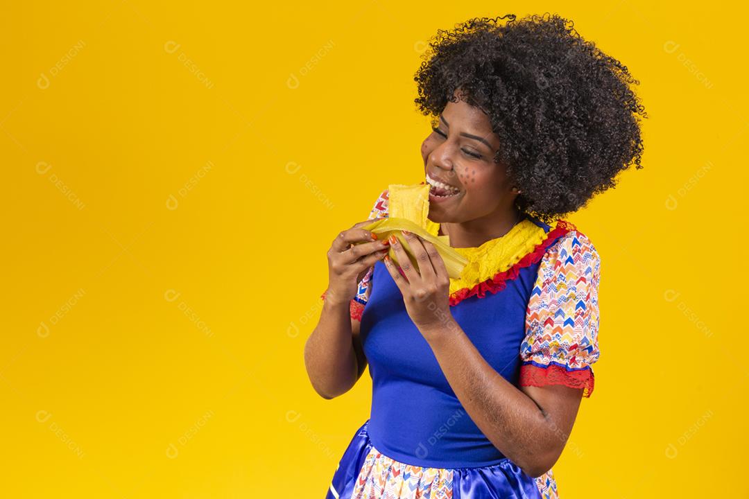 Mulher negra com roupas típicas da festa junina comendo uma deliciosa pamonha