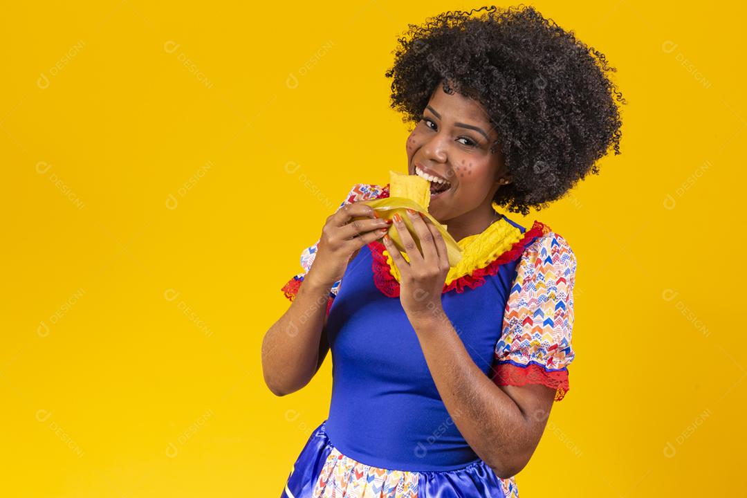 Mulher negra com roupas típicas da festa junina comendo uma deliciosa pamonha