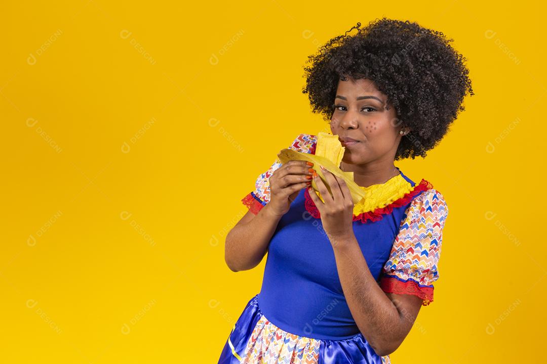 Mulher negra com roupas típicas da festa junina comendo uma deliciosa pamonha