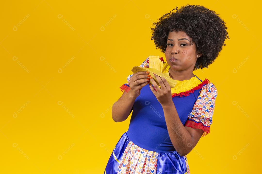 Mulher negra com roupas típicas da festa junina comendo uma deliciosa pamonha