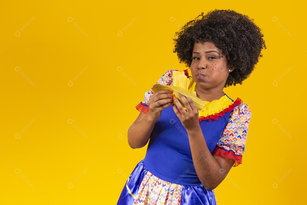 Mulher negra com roupas típicas da festa junina comendo uma deliciosa pamonha