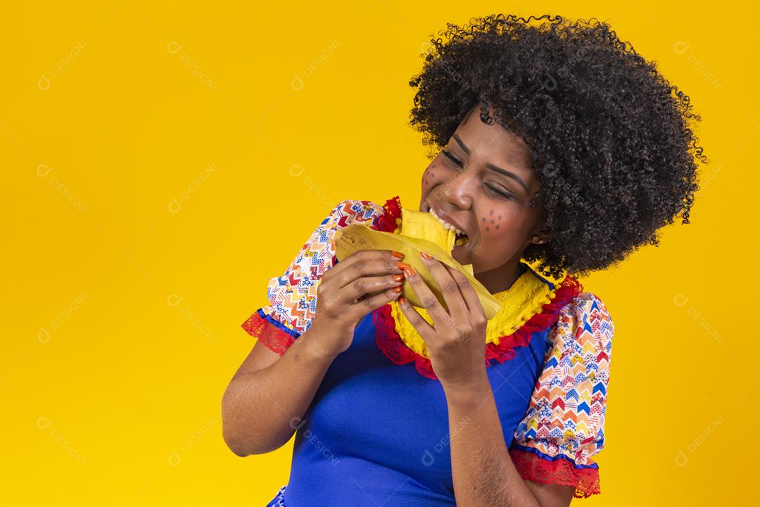 Mulher negra com roupas típicas da festa junina comendo uma deliciosa pamonha