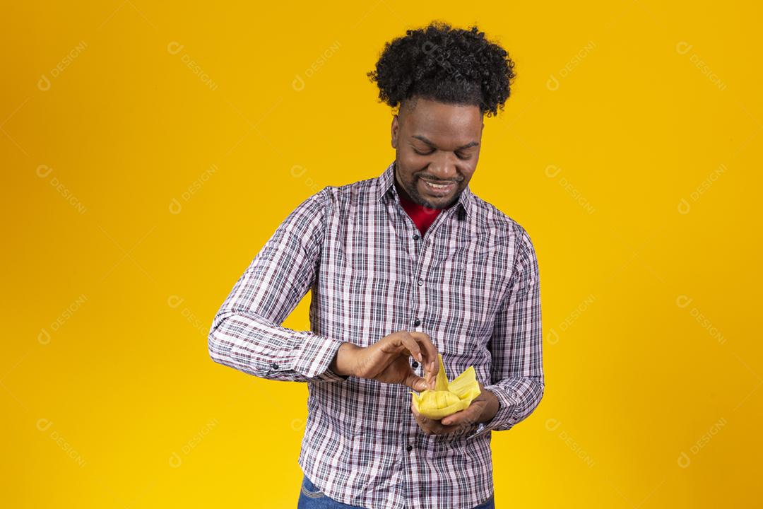 Homem negro com roupas típicas da festa junina comendo delicioso pamonha