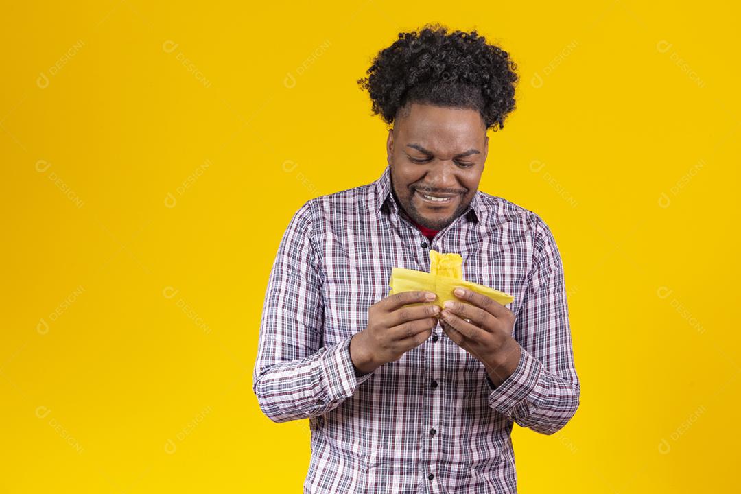 Homem negro com roupas típicas da festa junina comendo delicioso pamonha