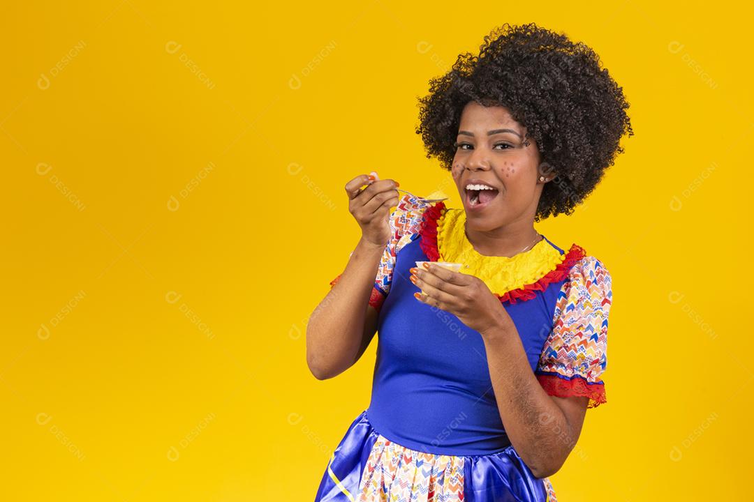 Mulher afro vestida com roupa de festa junina comendo um delicioso cural