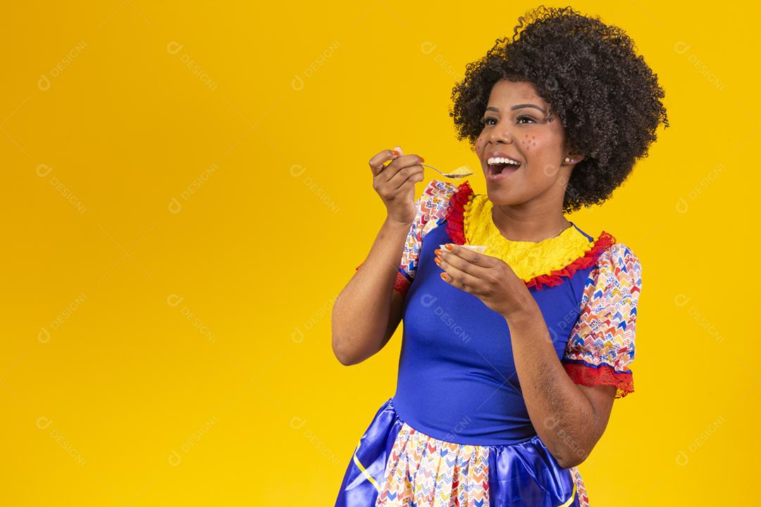 Mulher afro vestida com roupa de festa junina comendo um delicioso cural