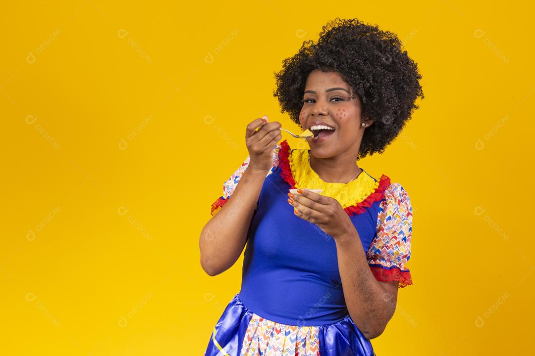 Mulher afro vestida com roupa de festa junina comendo um delicioso cural