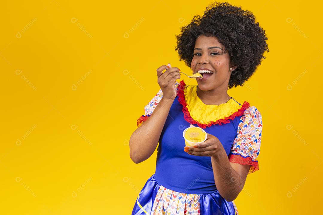 Mulher afro vestida com roupa de festa junina comendo um delicioso cural