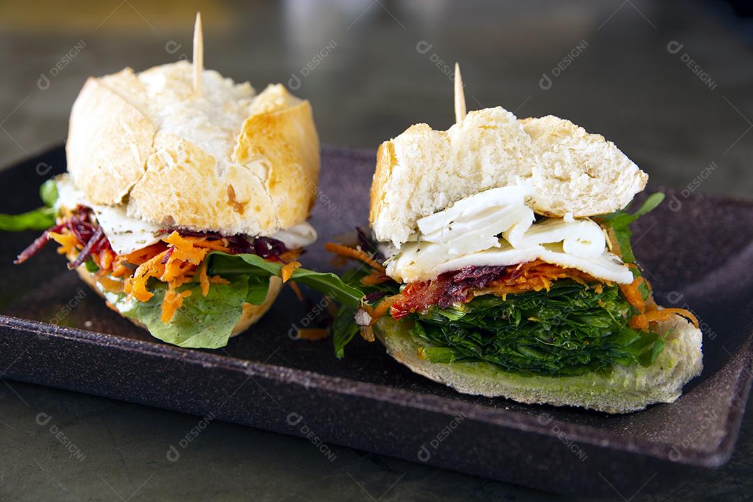 Sanduíche de queijo, cenoura, beterraba e alface em um prato preto sobre fundo de pedra.