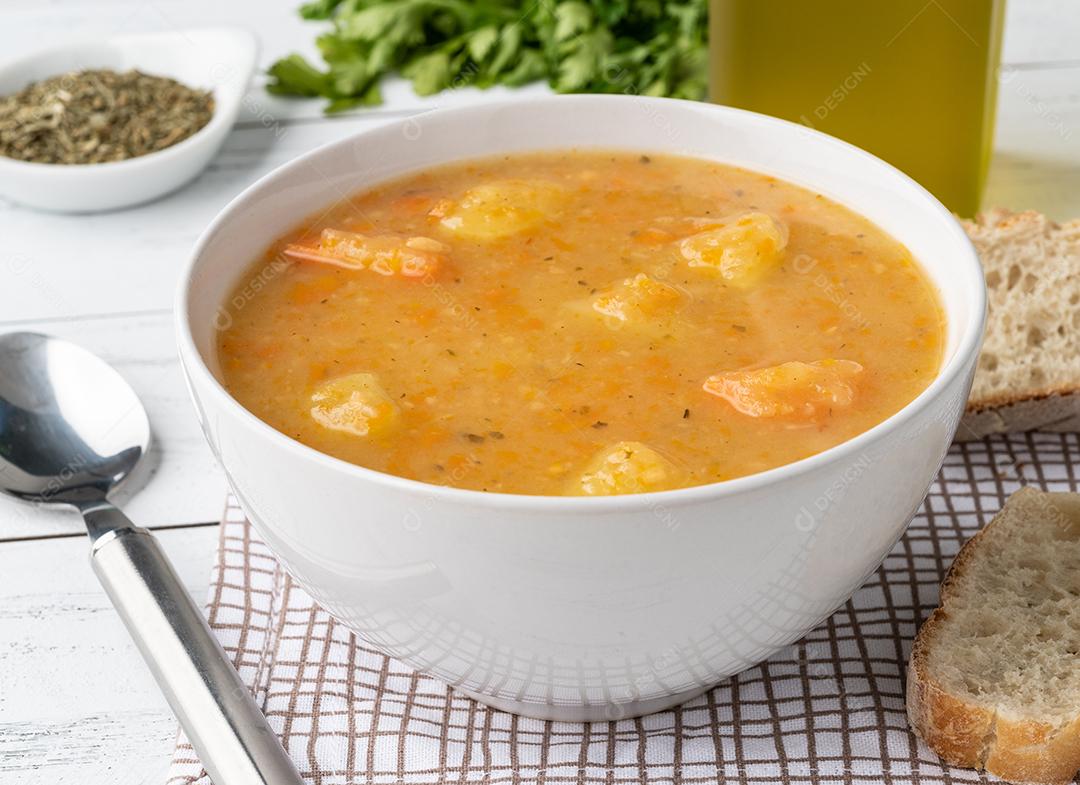 Sopa de legumes em uma tigela com temperos e fatias de pão sobre a mesa de madeira.