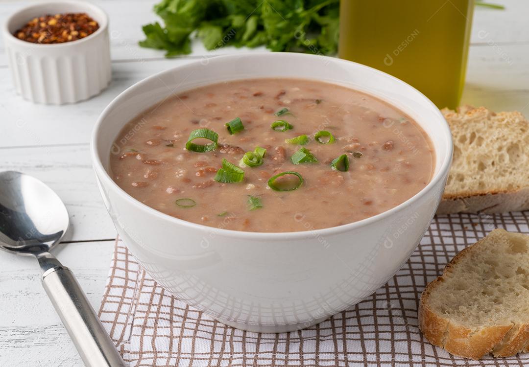 Sopa de feijão em uma tigela com fatias de pão e temperos sobre a mesa de madeira.