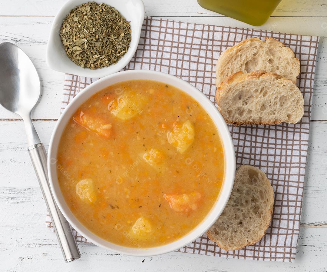 Sopa de legumes em uma tigela com temperos e fatias de pão sobre a mesa de madeira.