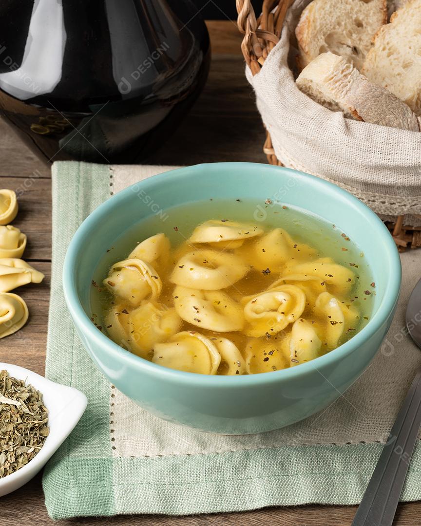 Cappelletti vs tortellini brodo sopa em uma tigela com fatias de pão e temperos sobre a mesa de madeira.