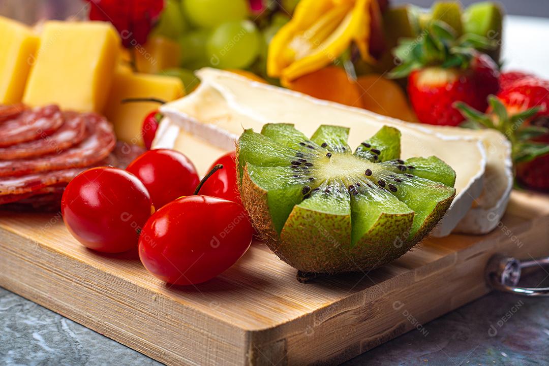 Tábua de frios e frutas servida em fundo de madeira e mármore com queijo, presunto, salame marrom, morangos, kiwis