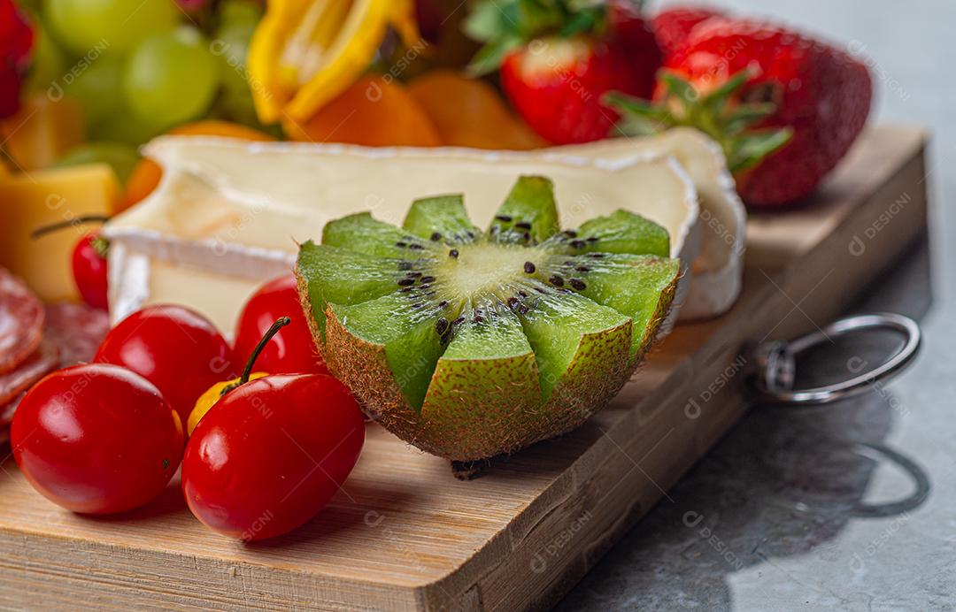 Tábua de frios e frutas servida em fundo de madeira e mármore com queijo, presunto, salame marrom, morangos, kiwis