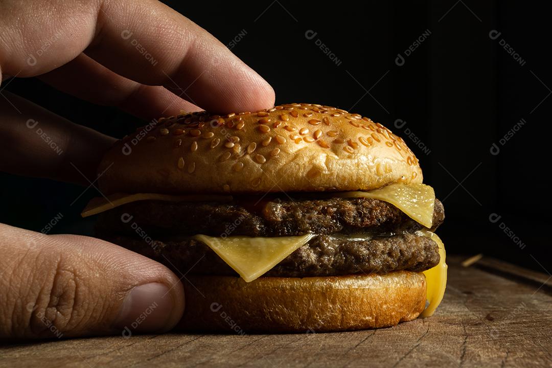 Hambúrguer ,carne grelhada, queijo com mão masculino segurando sobre mesa madeira fundo preto