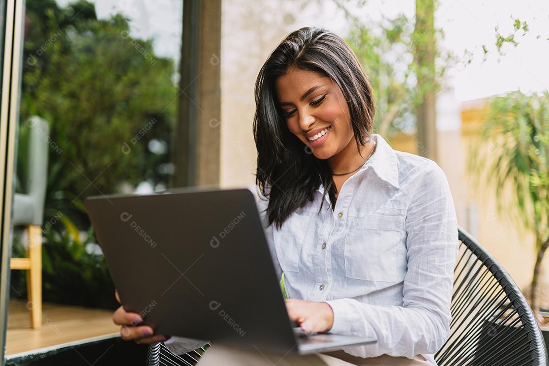 Nômade no trabalho. Confiante jovem latina fazendo videochamada no laptop enquanto está sentado pela janela no café.