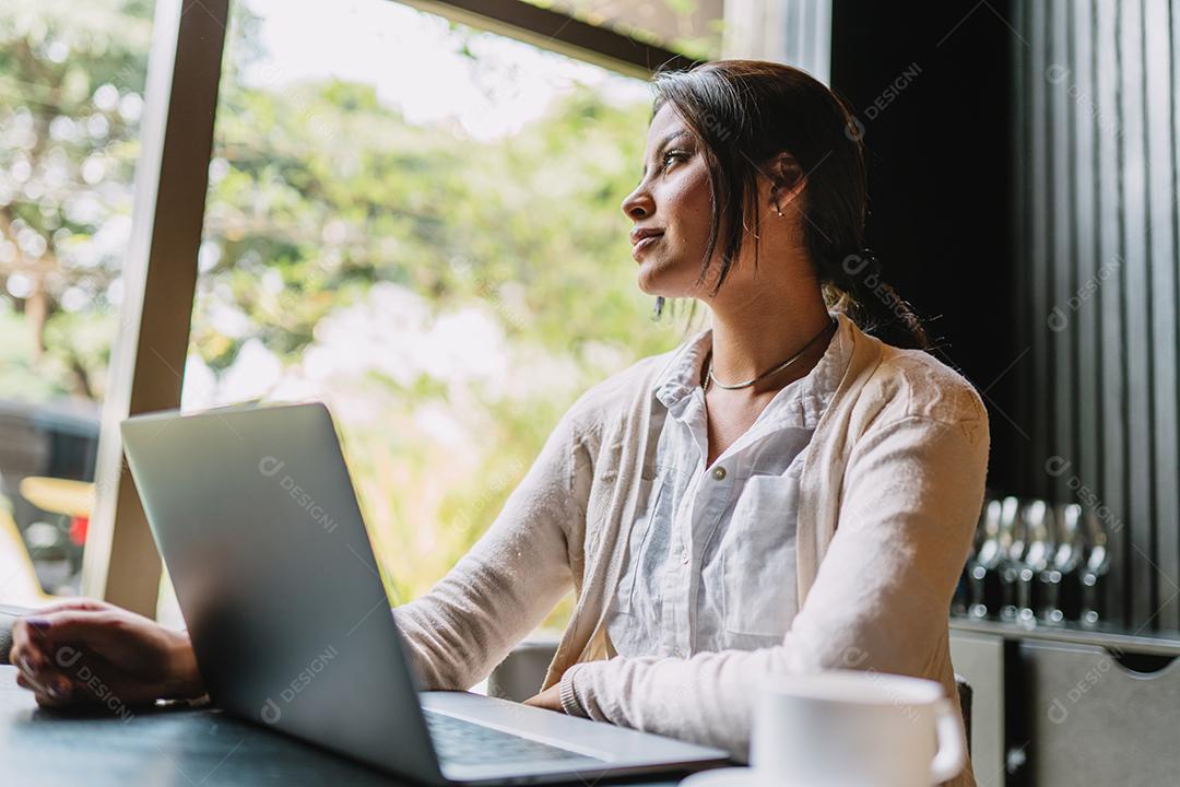 Nômade no trabalho. Confiante jovem latina fazendo videochamada no laptop enquanto está sentado pela janela no café.