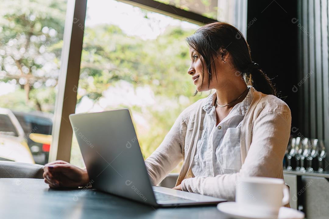 Nômade no trabalho. Confiante jovem latina fazendo videochamada no laptop enquanto está sentado pela janela no café.