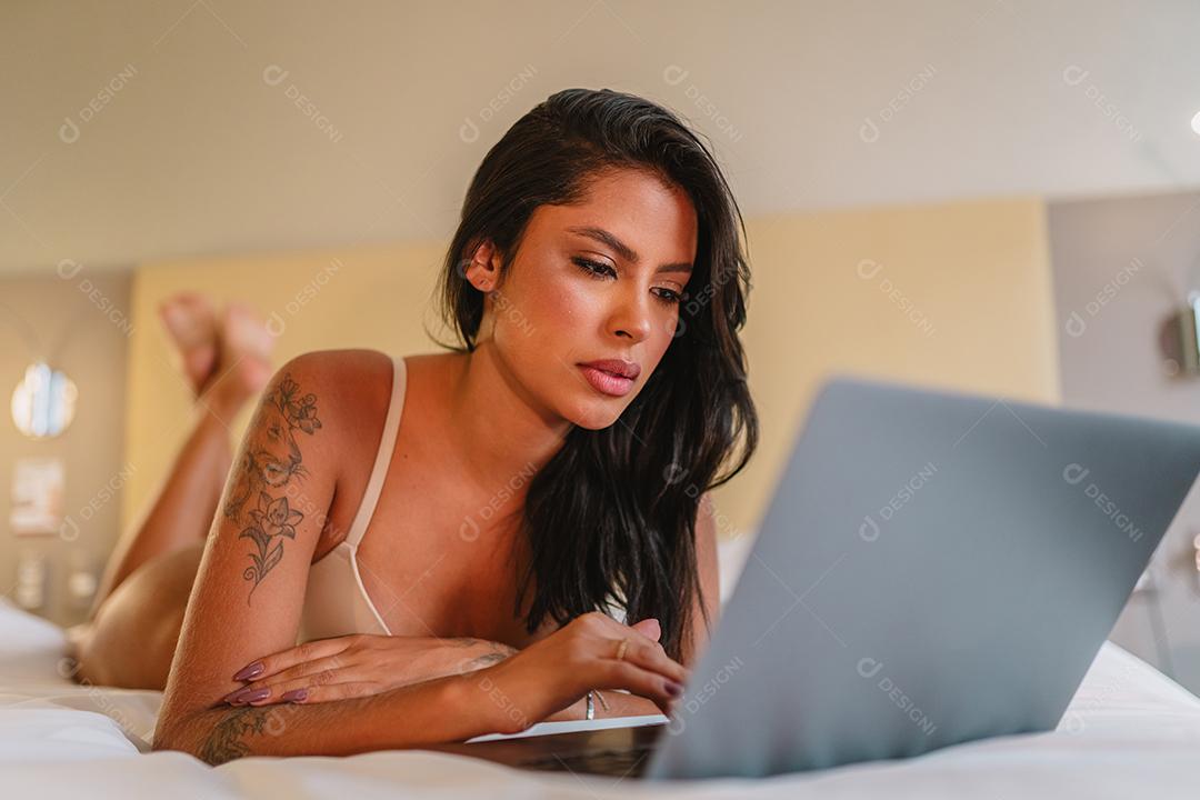 Mulher latina de 20 anos vestindo lingerie usando laptop enquanto estava deitada na cama no apartamento de hotel moderno