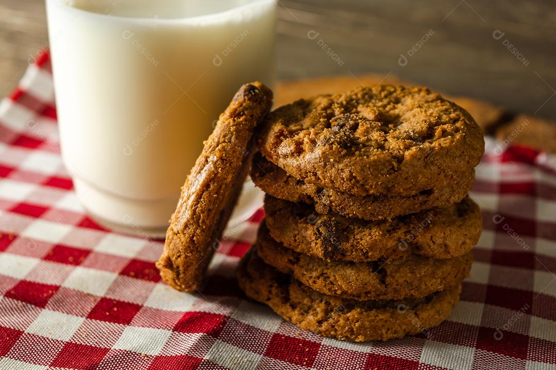 alguns biscoitos caseiros com gotas de chocolate, empilhados uns sobre os outros, e um copo de leite. na mesa de pano vermelho e fundo de madeira. conceito de café da manhã quente