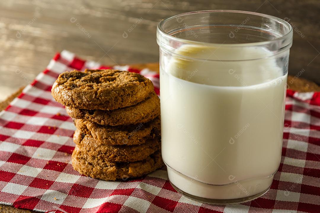 alguns biscoitos caseiros com gotas de chocolate, empilhados uns sobre os outros, e um copo de leite. na mesa de pano vermelho e fundo de madeira. conceito de café da manhã quente