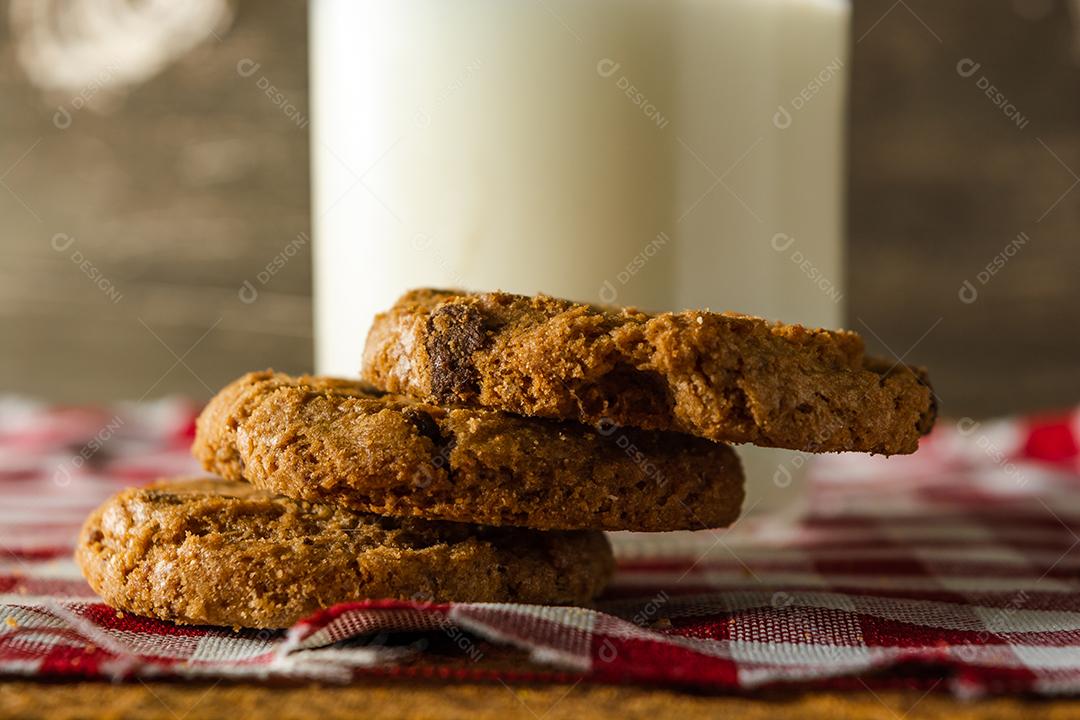 alguns biscoitos caseiros com gotas de chocolate, empilhados uns sobre os outros, e um copo de leite. na mesa de pano vermelho e fundo de madeira. conceito de café da manhã quente