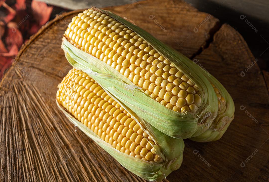 três deliciosos milhos amarelos crus com folha verde e espiga, sobre canudos secos e superfície de madeira