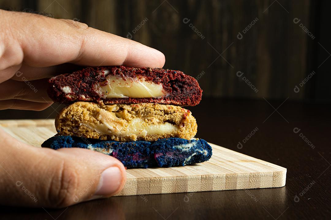 mão masculina apertando três biscoitos caseiros coloridos cortados ao meio em cima de uma superfície de madeira