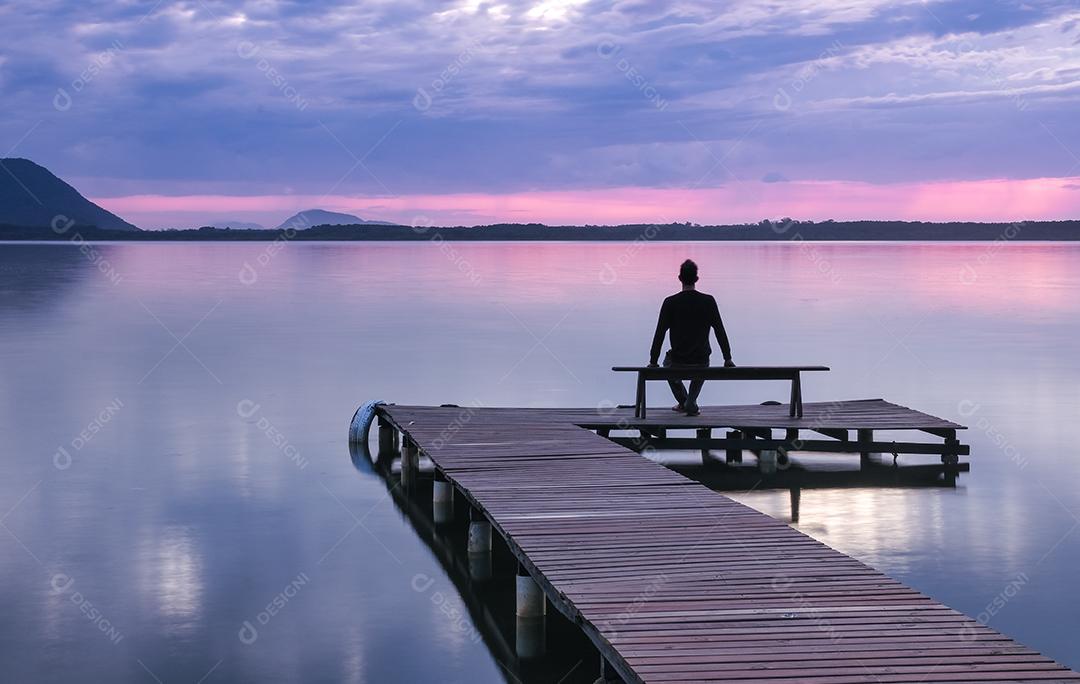 Homem sentado em um banco de cais de madeira assistindo o nascer do sol na Lagoa da Conceição em Florianópolis - Brasil