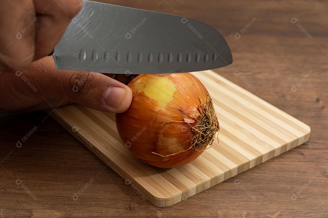 Mão segurando faca cortando cebola sobre tabua e mesa de madeira, tempero.