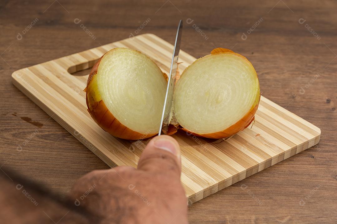 Faca cortando cebola ao meio sobre tabua e mesa de madeira, tempero.