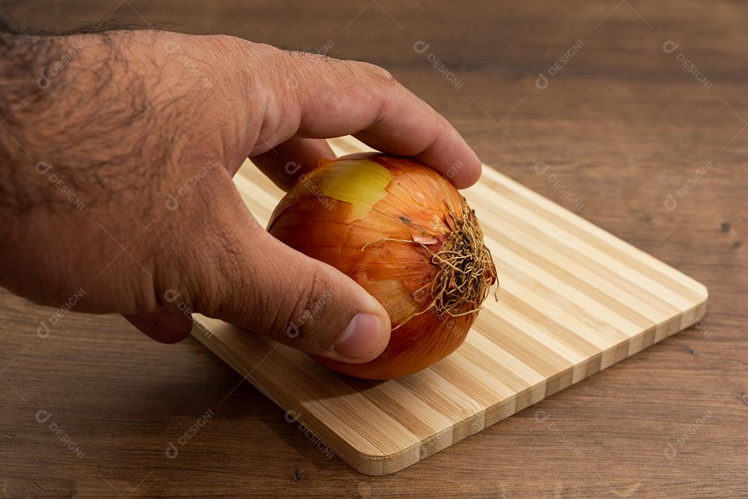 Mão segurando cebola sobre tabua e mesa de madeira, tempero.