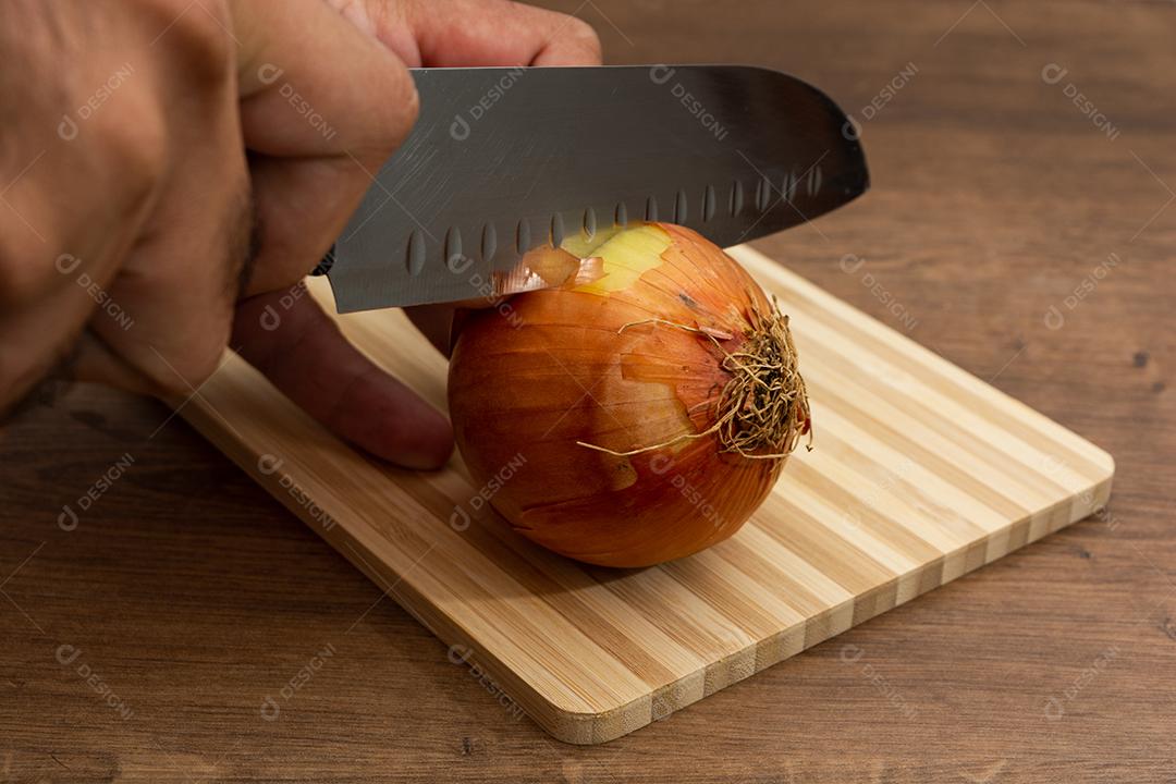 Faca cortando cebola ao meio sobre tabua e mesa de madeira, tempero.