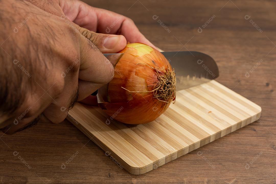 Faca cortando cebola ao meio sobre tabua e mesa de madeira, tempero.