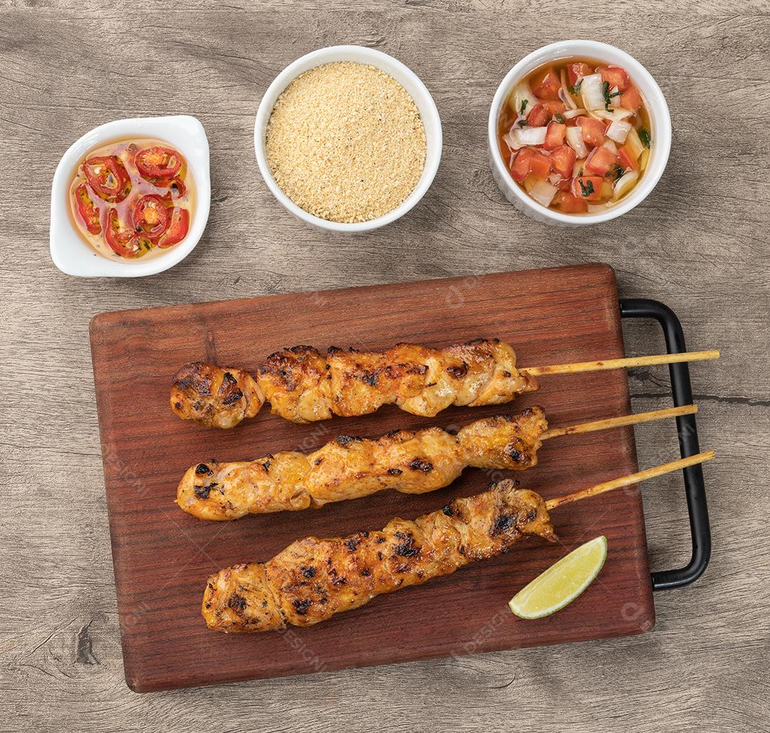 Espetinhos de frango na tábua de madeira com farofa e vinagrete.
