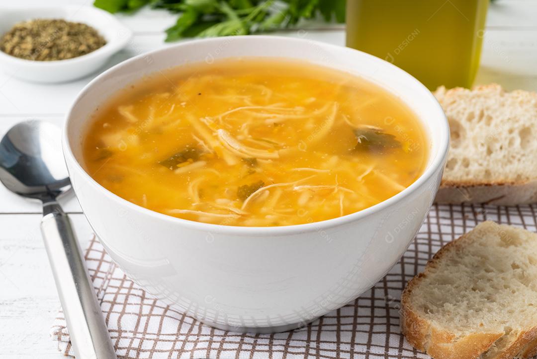 Sopa de galinha em uma tigela com fatias de pão e temperos sobre a mesa de madeira.