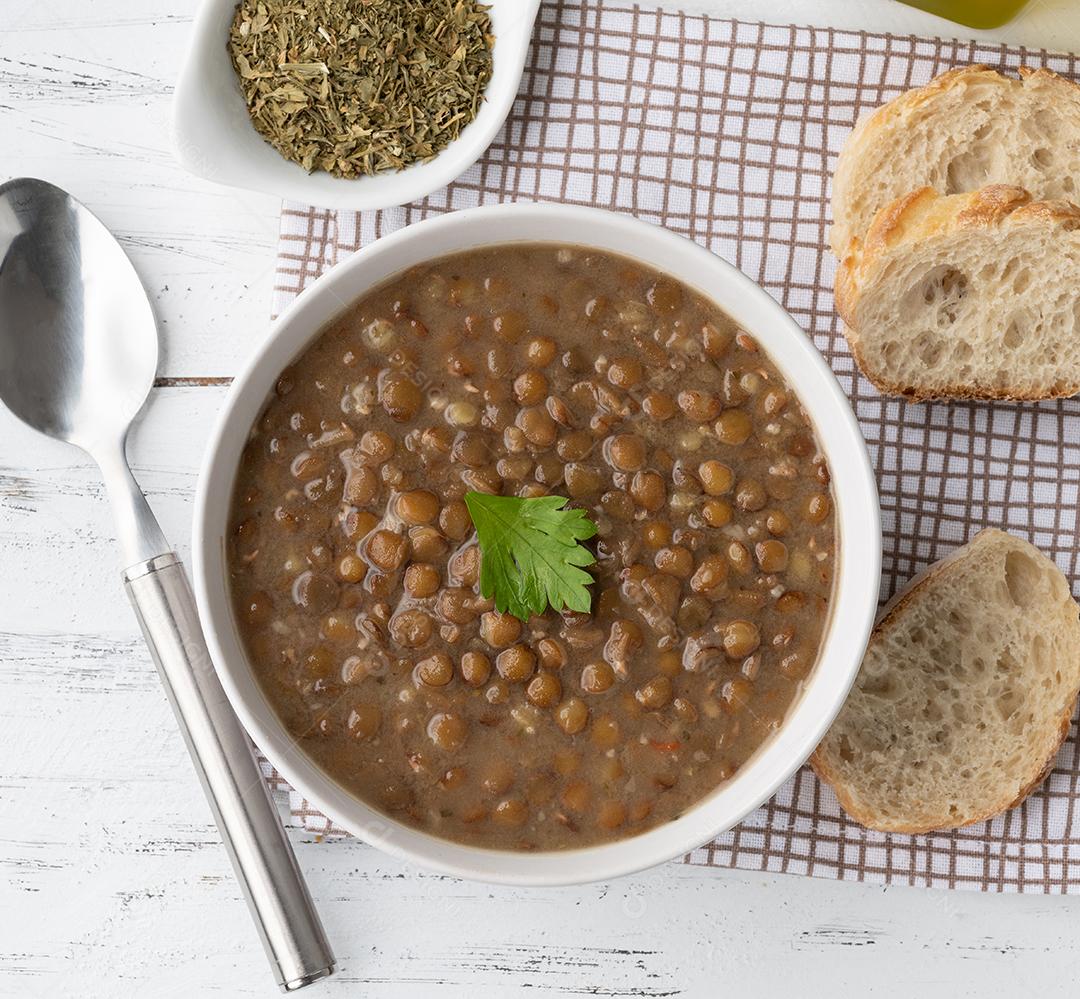 Sopa de lentilha marrom em uma tigela com fatias de pão e temperos sobre a mesa de madeira.