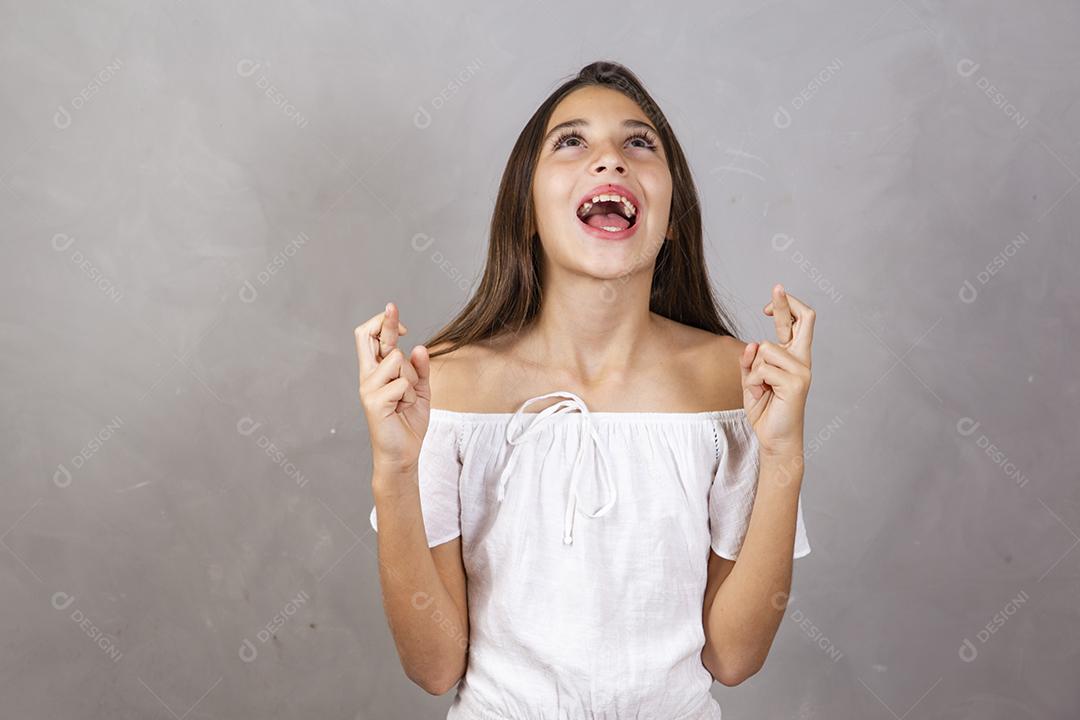 Menina pré-adolescente cruzando os dedos, esperando que seu desejo se torne realidade, isolado no fundo cinza do estúdio.