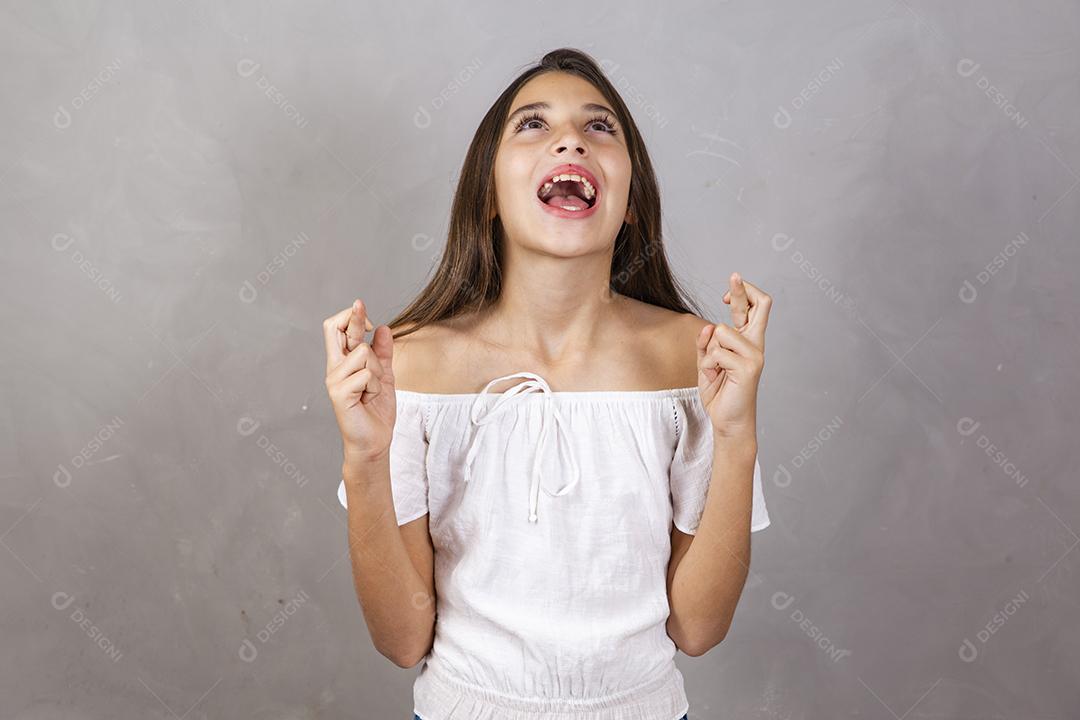 Menina pré-adolescente cruzando os dedos, esperando que seu desejo se torne realidade, isolado no fundo cinza do estúdio.