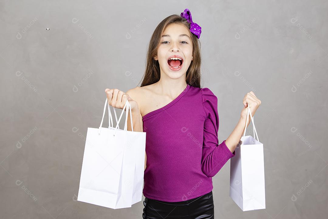 Menina segurando sacos de compras em pé isolado sobre fundo cinza. Moça de cerca de dez anos com sacolas de compras