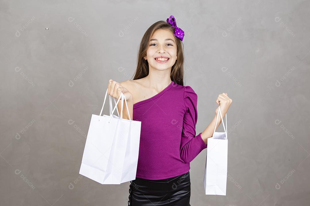 Menina segurando sacos de compras em pé isolado sobre fundo cinza. Moça de cerca de dez anos com sacolas de compras